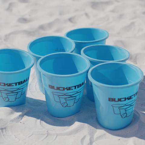 Six blue plastic cups with 'BUCKETBALL' logo on sand.
