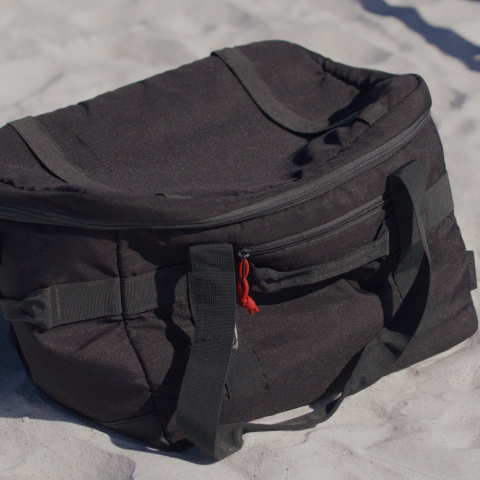 Black duffel bag with handles on white sand under sunlight.