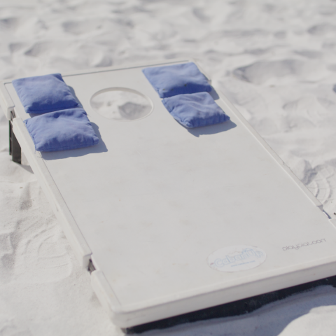 Cornhole board with blue bean bags on white sand beach.