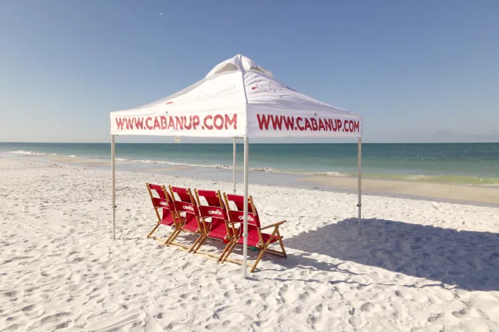 Four red chairs under a canopy on a sunny sandy beach with clear skies.
