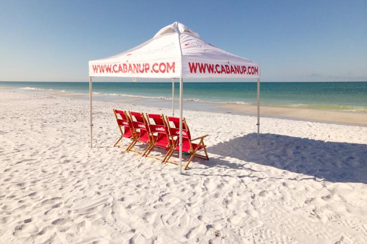 Four red chairs under a canopy on a sunny sandy beach with clear skies.
