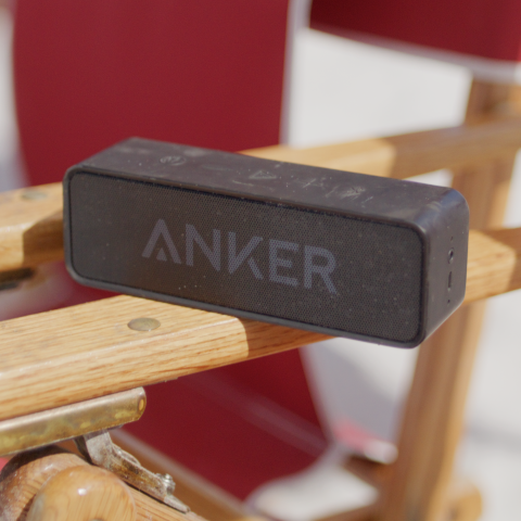 Black Anker speaker on wooden folding chair with red fabric in sunlight.